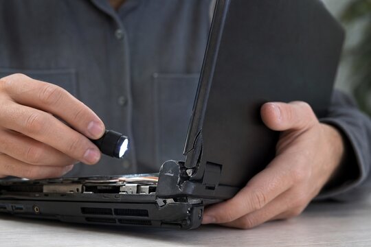 Close-up of person inspecting a damaged laptop hinge with a flashlight for repair