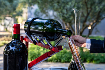 Obraz premium Professional sommelier pouring vintage red wine from a large format bottle into a glass decanter using a mechanical cradle holder for an outdoor luxury dining tasting event at a vineyard garden