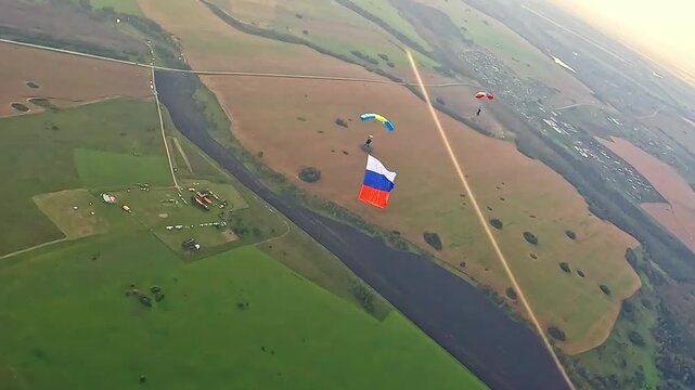 An aerial view of two parachutists over fields and forests. One athlete is descending with a large Russian tricolor.
