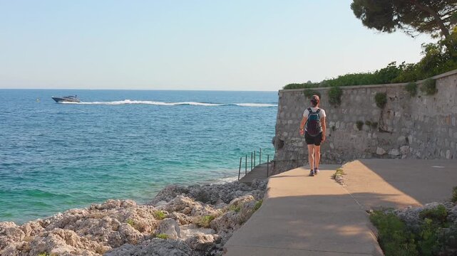Woman walks along the paved coastal Cap d'Ail hiking path above rocky shoreline as a motorboat speeds across the blue Mediterranean Sea, South of France