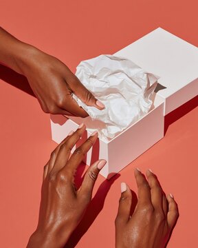 Dark skinned hands opening white gift box with crumpled tissue paper on coral background