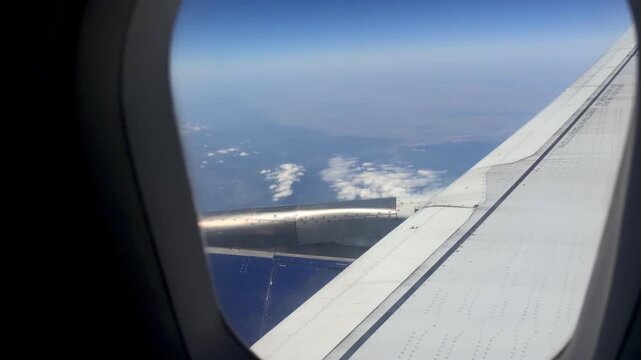 Airplane wing and engine visible through window, revealing clouds and blue sky while flying over a distant mountainous landscape