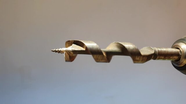 Close-up of a spiral auger drill bit spinning and rotating continuously, attached to a cordless power drill chuck against a neutral background.