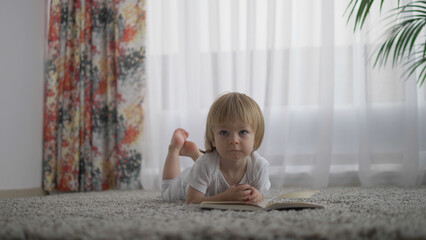 Fototapeta premium Baby little child lying down on carpet reading a book, play with feet, leisure, modern and traditional education