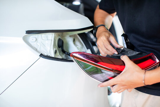 Mechanic hands installing and wiring a replacement tail light housing on a white vehicle in an auto repair shop.
