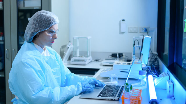 Female Scientist in Protective Gear Working on Laptop in Modern Laboratory with Blue Lighting
