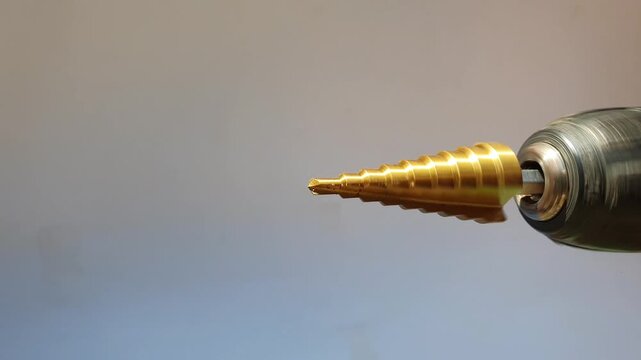Close up of a golden titanium-coated step drill bit spinning and rotating inside the chuck of a cordless electric hand drill against a neutral gradient background.