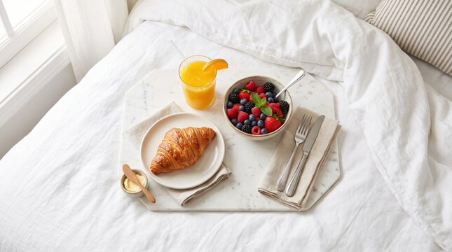 A cozy breakfast in bed setup featuring a croissant, a bowl of mixed berries with yogurt, and a glass of orange juice on a white tray.