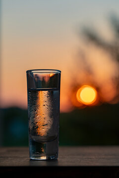 Cold shot glass with clear alcohol at sunset. Chilled shot glass filled with transparent alcohol or vodka covered in condensation drops stands on wooden table against blurry warm orange sunset