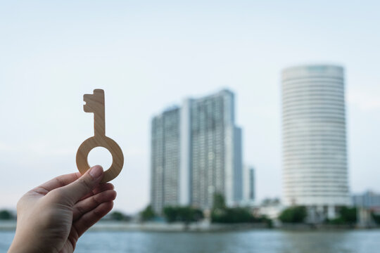 Real estate and urban property investment concept with hand holding wooden key against river and modern skyscraper condominium buildings for city apartment buying and home ownership.