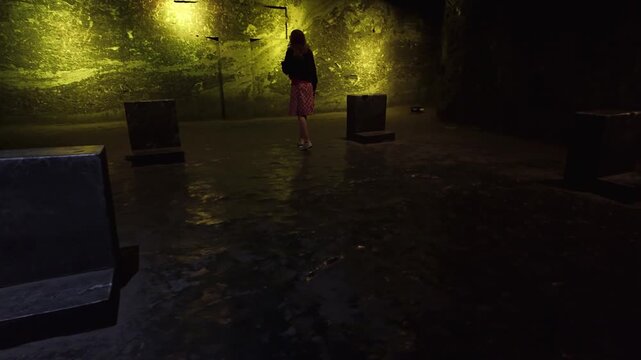 Woman listening to an audio guide while walking through the illuminated Salt Cathedral of Zipaquira, Colombia, approaching a stone cross carved into the wall, cultural heritage concept