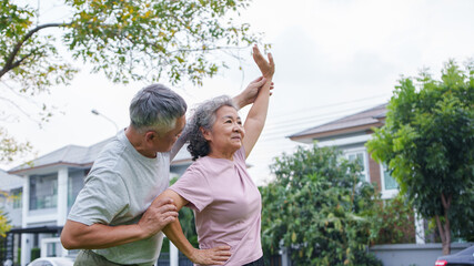 Senior Asian woman exercising outdoors with a husband supporting his wife during an arm stretch,...