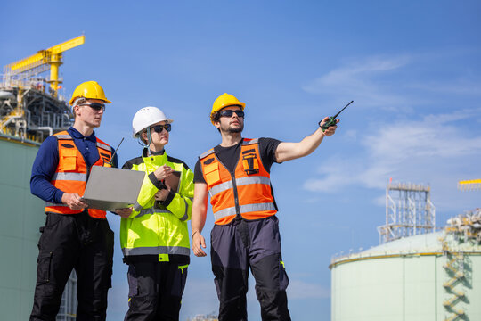 Industrial engineers team with laptop and walkie talkie at refinery plant, Diverse project manager and workers team in safety gear pointing and collaborating at industrial site under blue sky