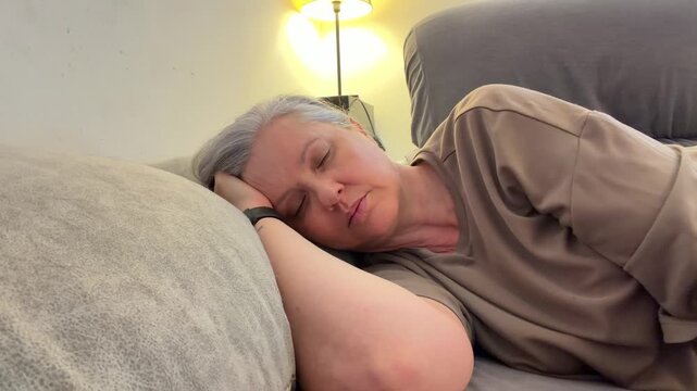 A woman lays on a couch in her living room with her head resting on her arm. She appears to be taking a nap under soft light. It is afternoon.