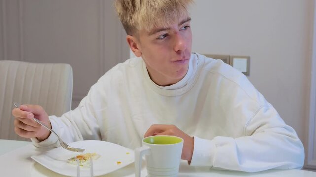 Young blonde man sits at dining table, feeling pensive and looking away during quiet meal at hom