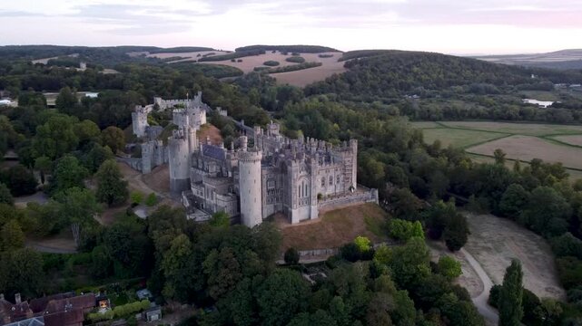 Slow cinematic drone orbit around historic Arundel Castle at sunrise, surrounded by trees and the rolling countryside of West Sussex, England