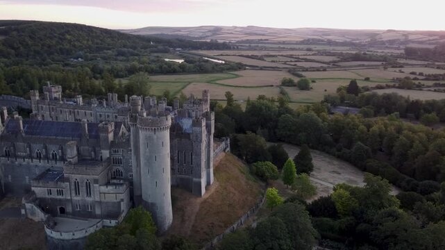 Cinematic high aerial orbit around Arundel Castle at sunrise, revealing the historic fortress, surrounding woodland and expansive West Sussex countryside in warm golden light