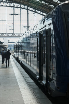 Passengers preparing to board a modern train at a historic railway station, framed by dramatic daylight through an expansive glass and steel roof.