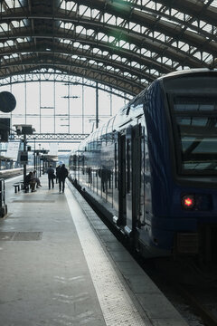 Passengers preparing to board a modern train at a historic railway station, framed by dramatic daylight through an expansive glass and steel roof.
