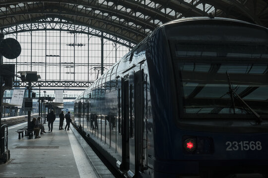 Passengers preparing to board a modern train at a historic railway station, framed by dramatic daylight through an expansive glass and steel roof.