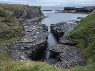 Rugged cliffs frame a narrow, dark tidal pool. Green grass lines the rocky edges. Water rushes between jagged stones. Overcast skies add a moody tone. Perfect for nature or travel themed projects © ANTONIO