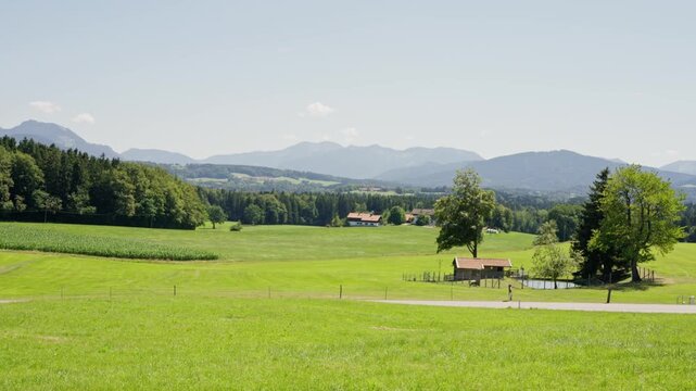 Green meadow panorama with farmhouse, pond, lone tree, and motorcycle passing on rural road under layered mountain ridgelines, with landscape, meadow, field, pasture detail