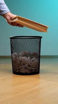Hand discarding an old book, symbolizing the end of knowledge, outdated information, or a finished project, into a black mesh trash bin filled with crumpled paper