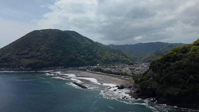 Sand beach lined with concrete breakwaters below a forested mountain and coastal town in Kawazu, Izu, Japan, on an overcast day, aerial drone shot