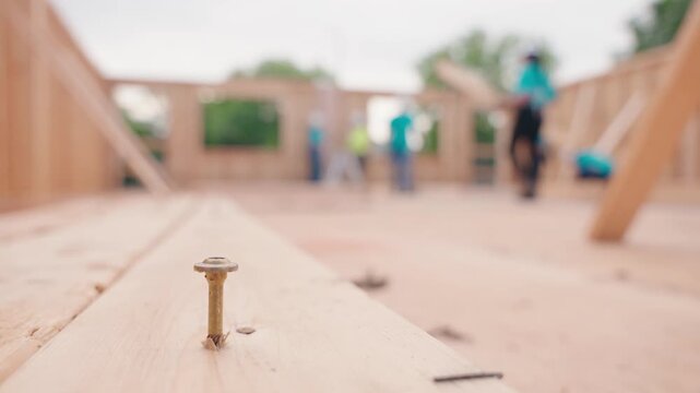 Static shot with a construction screw in the foreground as the camera slowly zooms toward home builders working in the background inside a partially constructed house