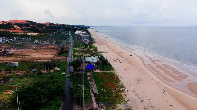 People walk along Jumping Rocks Beach in Tan Thanh, Lam Dong. The shore stretches with buildings and nature in view. Waves hit the sand while clouds drift above.