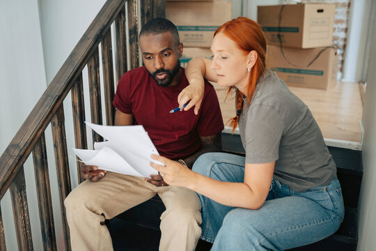 Diverse couple going through paperwork while sitting on staircase in new apartment