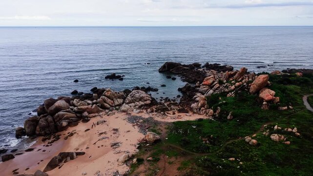 Visitors walk along Jumping Rocks Beach in Tan Thanh, Lam Dong. The clear blue water meets the sandy shore. Large rocks dot the landscape while people enjoy the beach.
