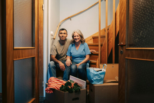 Portrait of smiling couple sitting on wooden stairs surrounded by moving boxes
