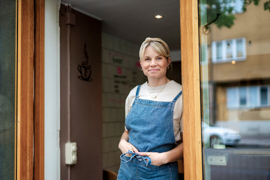 Portrait of female shop owner with hands in pockets leaning against wooden door frame