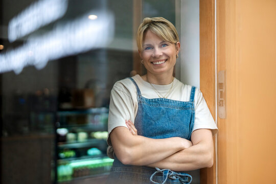 Portrait of smiling female owner posing with arms crossed while leaning on glass outside shop
