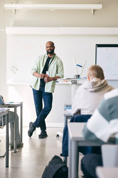 Full length of male mathematics professional leaning on desk while teaching students in high school classroom