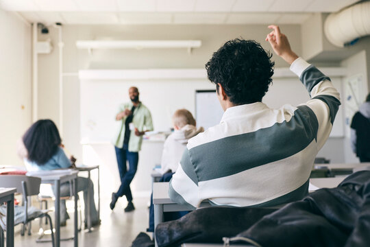 Rear view of teenage boy raising hand while asking question during college lecture