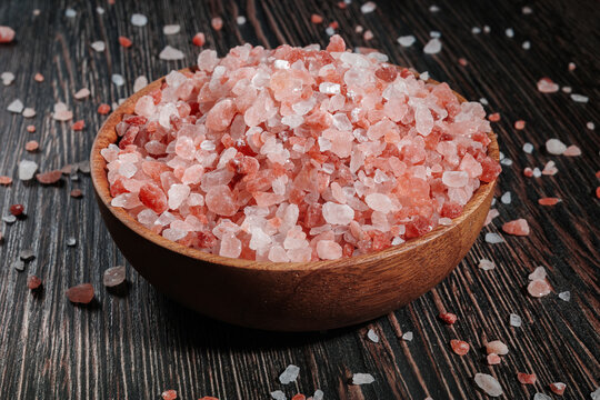 Wooden bowl filled with pink Himalayan salt crystals on dark wooden table, natural mineral seasoning for healthy cooking and spa concepts.