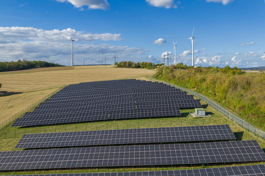 Aerial view of Photovoltaik-Anlage Wallrabenstein featuring rows of solar panels in a rural landscape with wind turbines in the background in Hunstetten, Hessen, Germany.