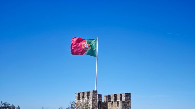 Flag of Portugal waving atop a stone castle tower against a clear blue sky, showcasing historical architecture and national pride in a scenic outdoor setting