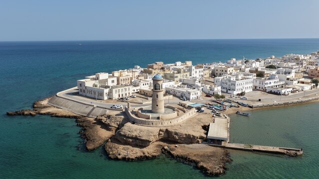Aerial view of the historic Al Ayjah Lighthouse on a rocky promontory overlooking the coastal town of Sur with white buildings and turquoise waters in Sur, Ash Sharqiyah South G, Oman.