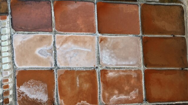 Aerial view of salt pans in Qurayyat, showing the grid of rectangular evaporation ponds with varying shades of brown, orange, and white salt deposits in Qurayyat, Muscat Governorate, Oman.