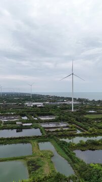 An aerial view of wind turbines for electricity generation near the sea with shrimp ponds and farmland below captured by a drone camera.