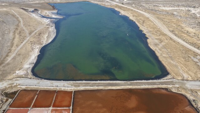 Aerial view of the salt pans and lagoon, featuring vibrant red and green water pools separated by dirt paths in the arid landscape of Qurayyat, Muscat Governorate, Oman.