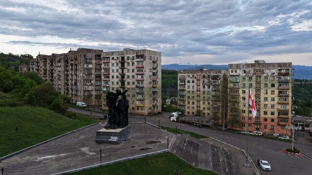 Aerial view of Soviet-era apartment blocks and a central monument in a plaza with the flag of Georgia and mountains in the background in Chiatura, Imereti, Georgia.