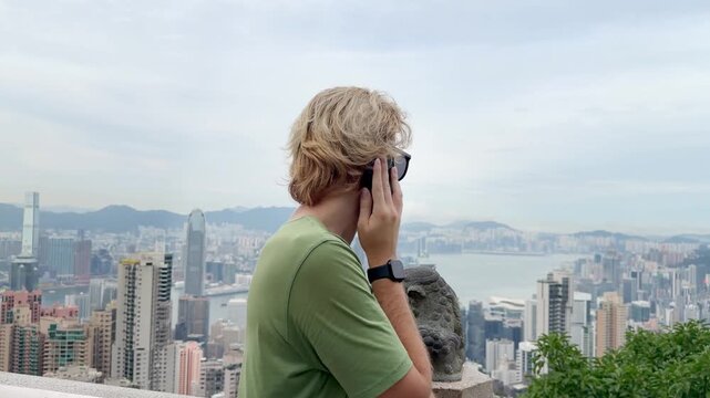 Young Blond Man Using Smartphone and Photographing with View of Hong Kong Skyscrapers from Above &ndash; Urban Panorama with City Skyline