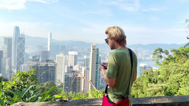 Young Blond Man Using Smartphone and Photographing with View of Hong Kong Skyscrapers from Above &ndash; Urban Panorama with City Skyline