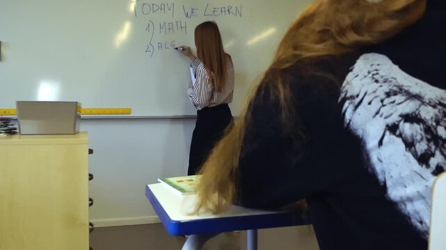 Young woman professor writing Today we learn algebra on a whiteboard while a student with long hair watches attentively from her desk during a math lesson inside a modern classroom