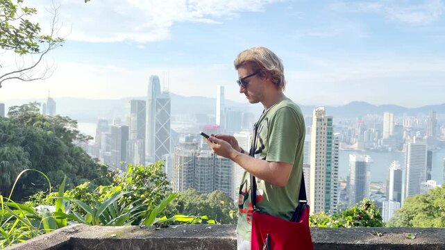 Young Blond Man Using Smartphone and Photographing with View of Hong Kong Skyscrapers from Above &ndash; Urban Panorama with City Skyline