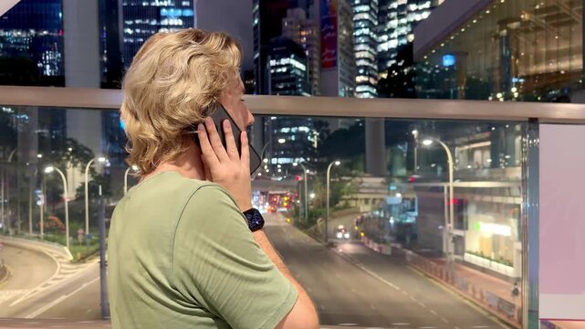 Young Blond Man Talking on Smartphone with Evening City Lights and Skyscrapers Background &ndash; Night Urban Scene